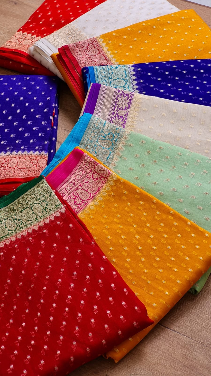 A collection of folded colorful fabric pieces, likely traditional sarees or dupattas, arranged diagonally on a light wooden floor. The fabrics feature white zari dot patterns and ornate borders in red, yellow, blue, mint green, pink, and white.