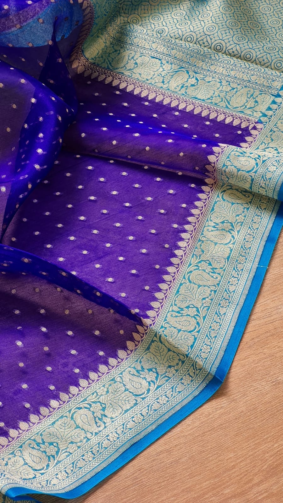 A close-up of a purple Banarasi silk saree with intricate light blue and white zari embroidery, shown draped on a wooden surface.