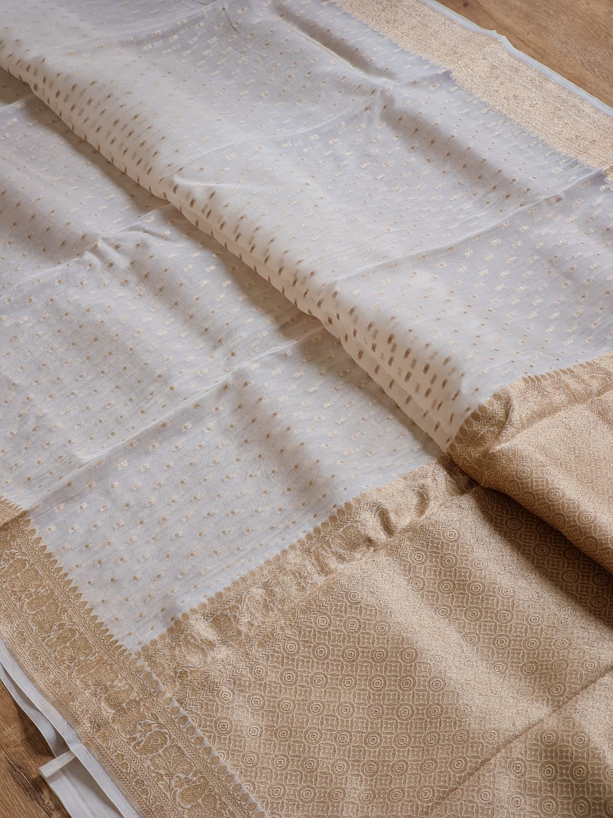 A close-up of a traditional Indian saree or fabric, featuring a white body with small gold zari motifs and a wide beige border with intricate woven patterns.