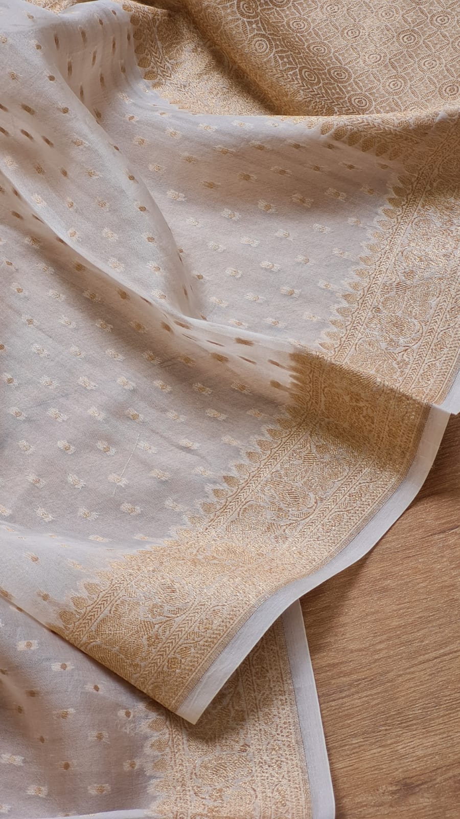 Light beige and off-white fabric with delicate floral embroidery and a contrasting golden-beige border, draped over a wooden surface.