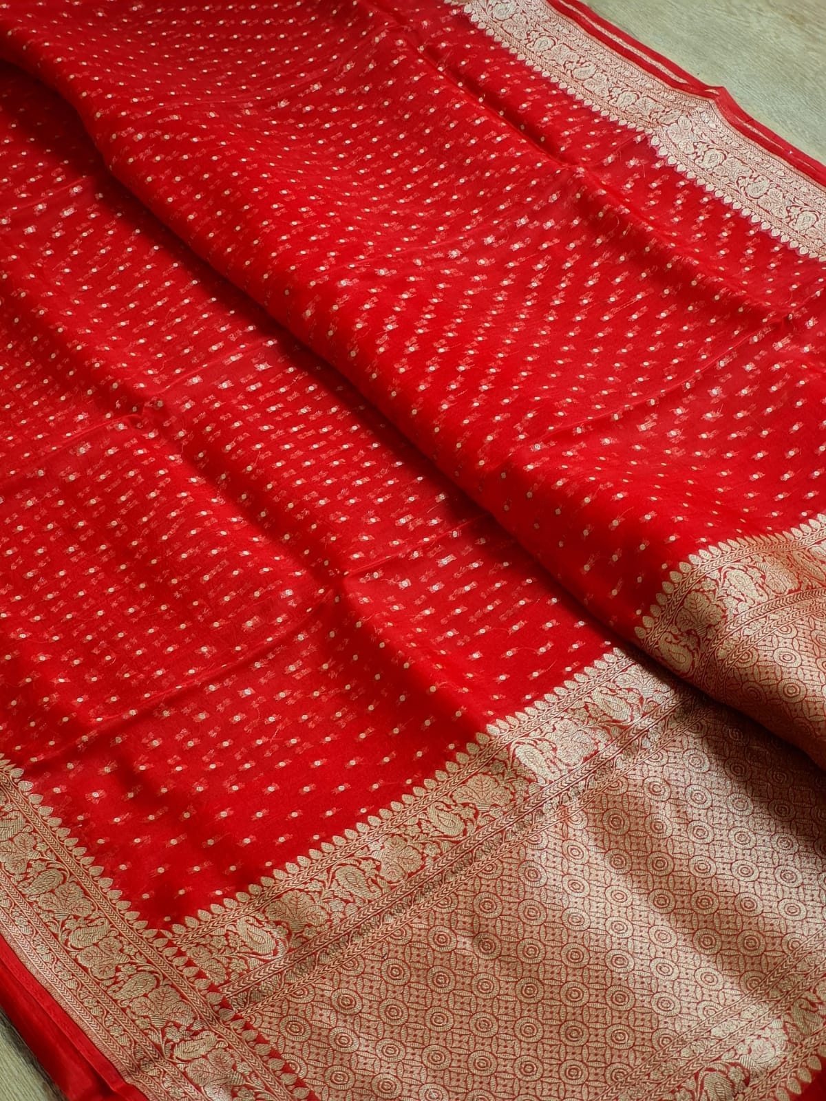 A close-up of a red fabric textile with a dense small white dot pattern and a wide beige-gold woven border.
