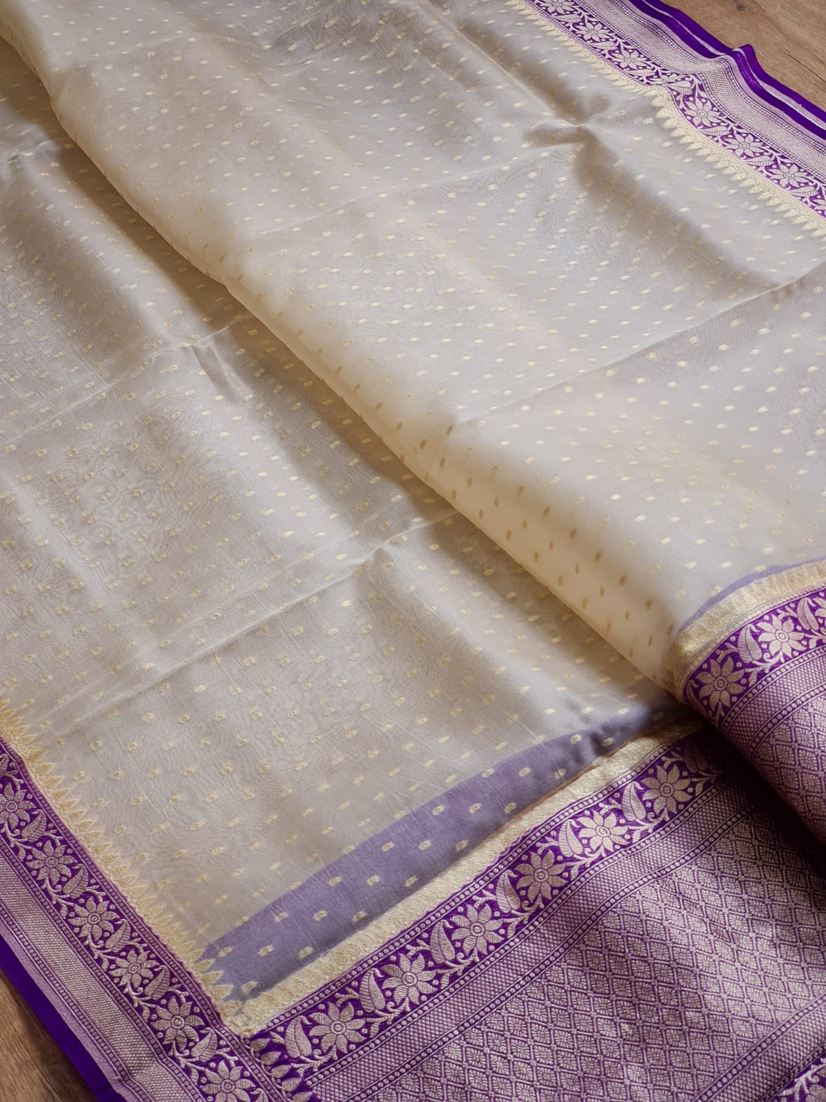 Ivory -colored silk saree with subtle gold zari embroidery, featuring a contrasting purple border with intricate floral patterns.