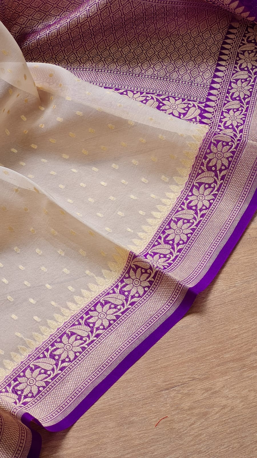 A close-up of a traditional Indian saree or fabric, featuring a cream-colored body with small gold motifs and a rich purple border with floral patterns, laid flat on a light wooden surface.