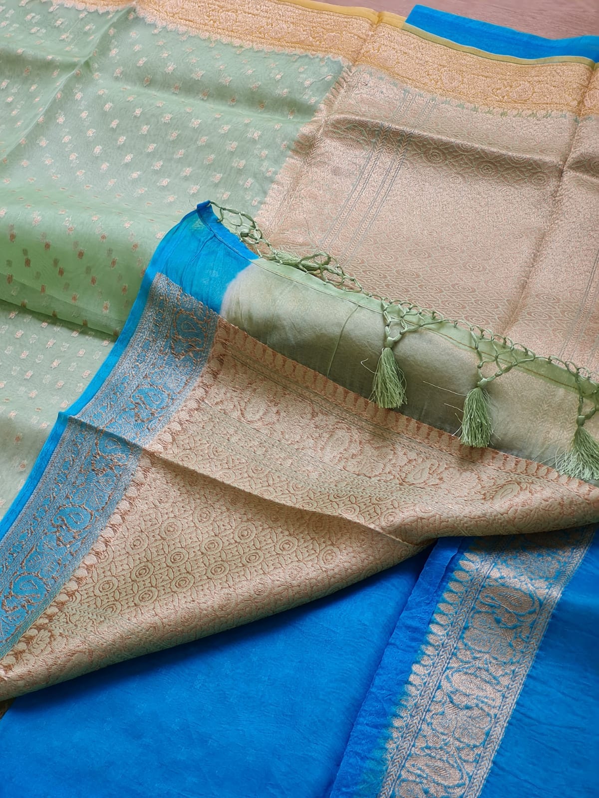 A folded and draped saree, showing a light sage green body with tiny gold motifs, a beige and gold zari border, and a vibrant royal blue pallu with intricate gold woven borders.