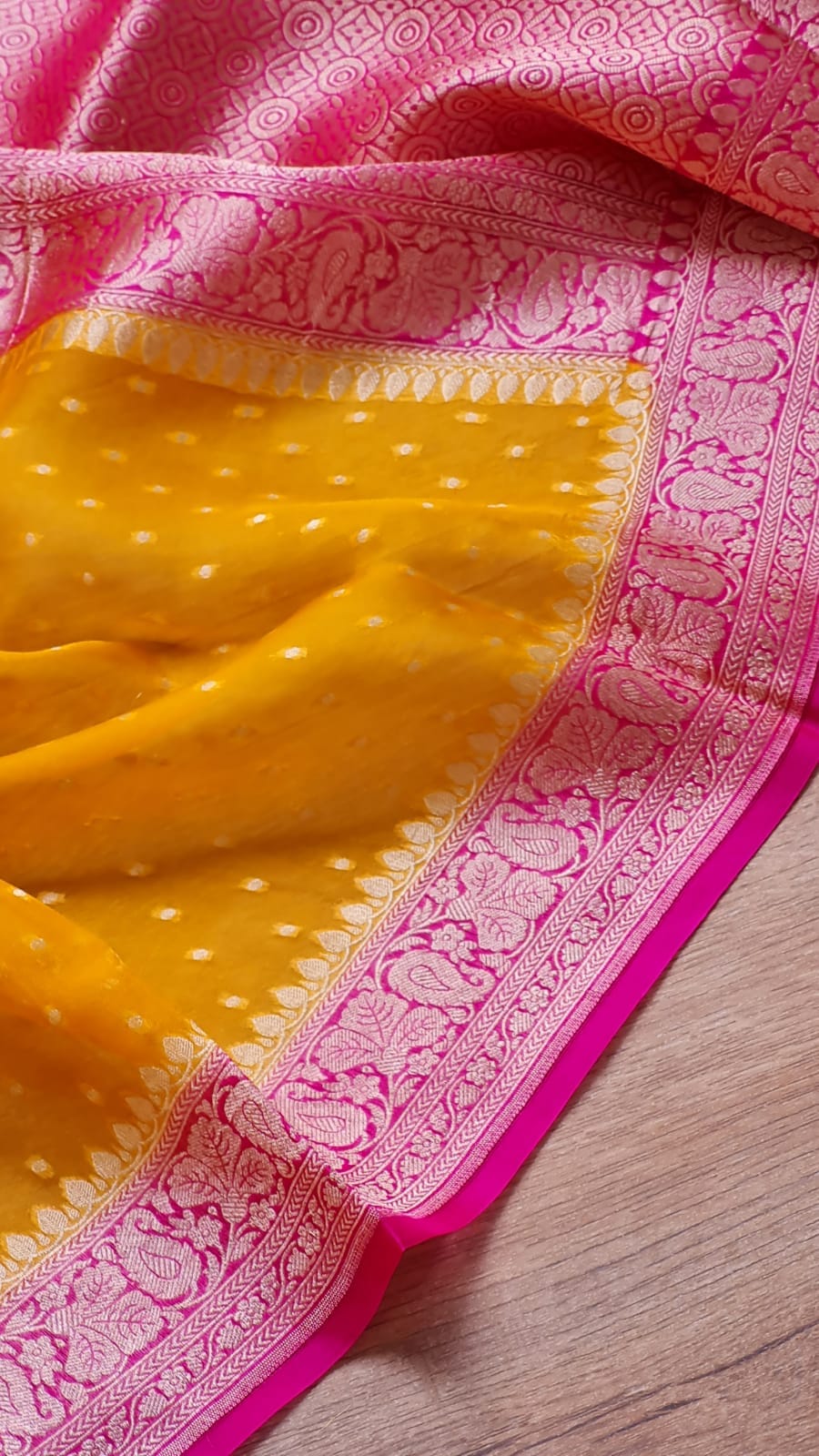 Yellow and pink Banarasi silk saree, showcasing its vibrant yellow body with small white dots, a rich pink border with intricate metallic zari embroidery, and a matching pink end piece draped on a light wood surface.