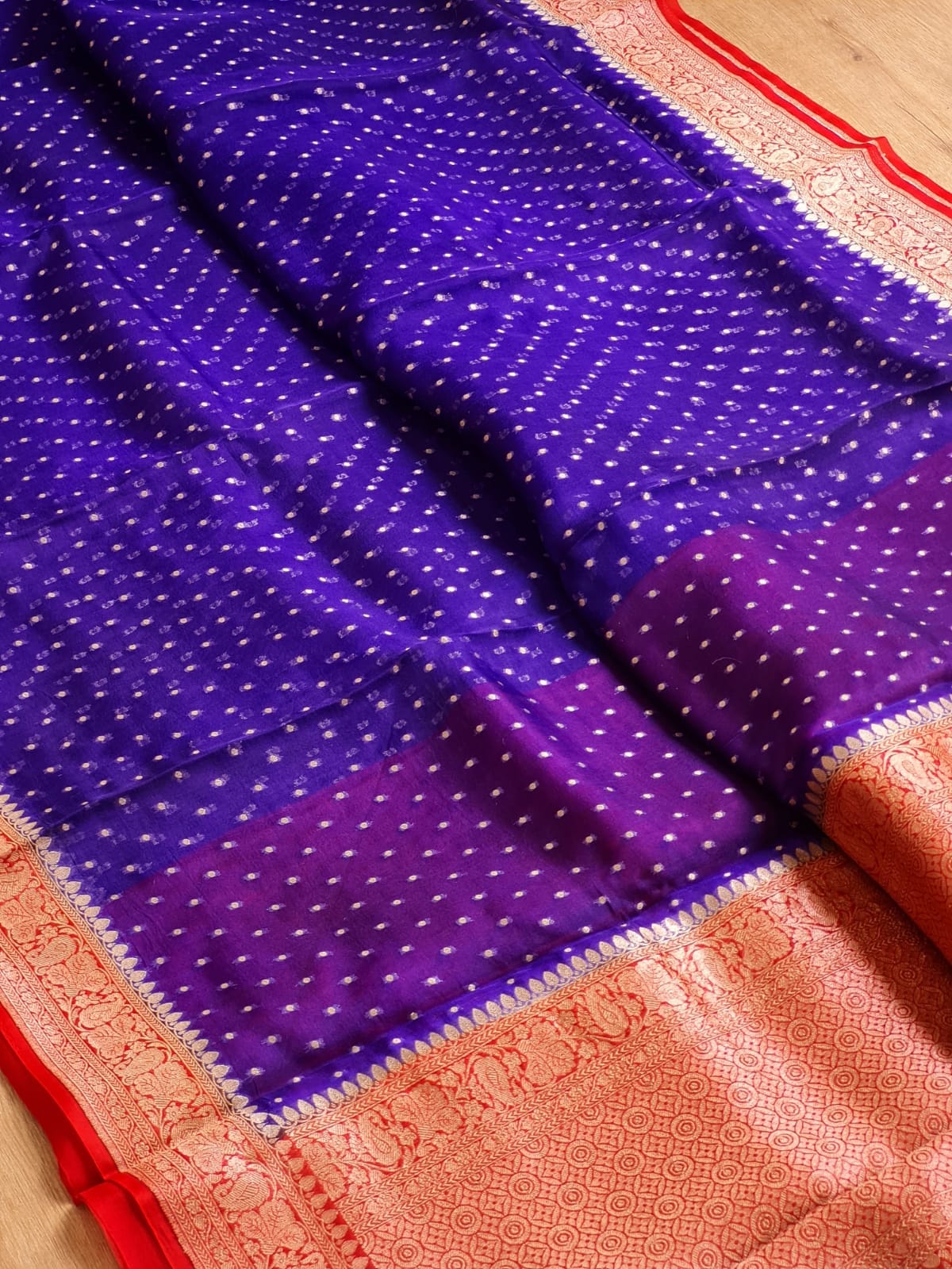 A close-up of a purple and magenta patterned fabric, likely a saree, featuring small white dot motifs and a wide peach-pink border with red accents.