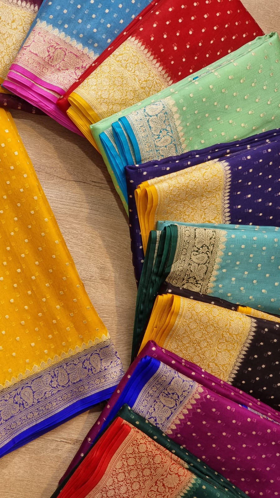 A collection of folded colorful sarees arranged in a fan shape on a light wooden surface. The sarees display a variety of vibrant colors including yellow, blue, red, green, purple, and orange, each with traditional patterns and borders.