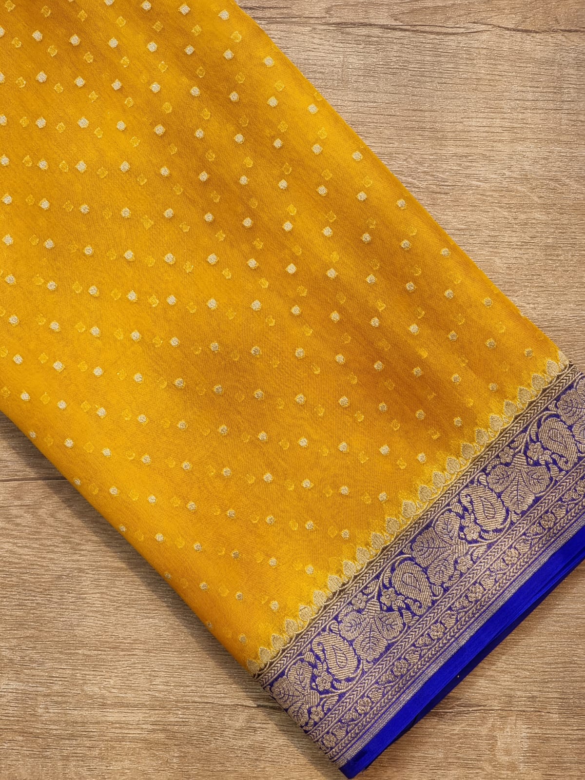 Yellow Banarasi silk saree fabric with small gold zari motifs and a detailed blue and gold border, laid flat on a wooden surface.