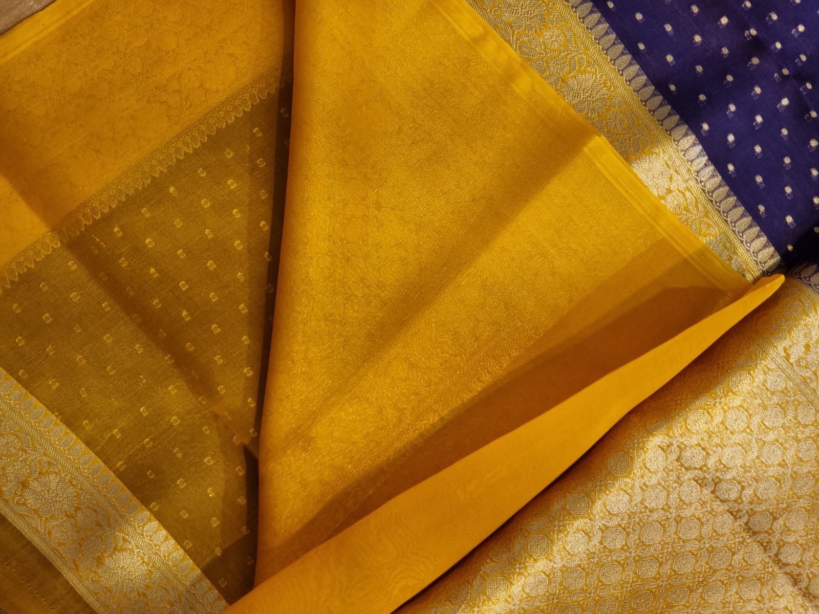 A folded yellow-gold fabric, likely a silk saree, showing its subtle sheen, dotted pattern, and a contrasting deep purple border with gold zari work.
