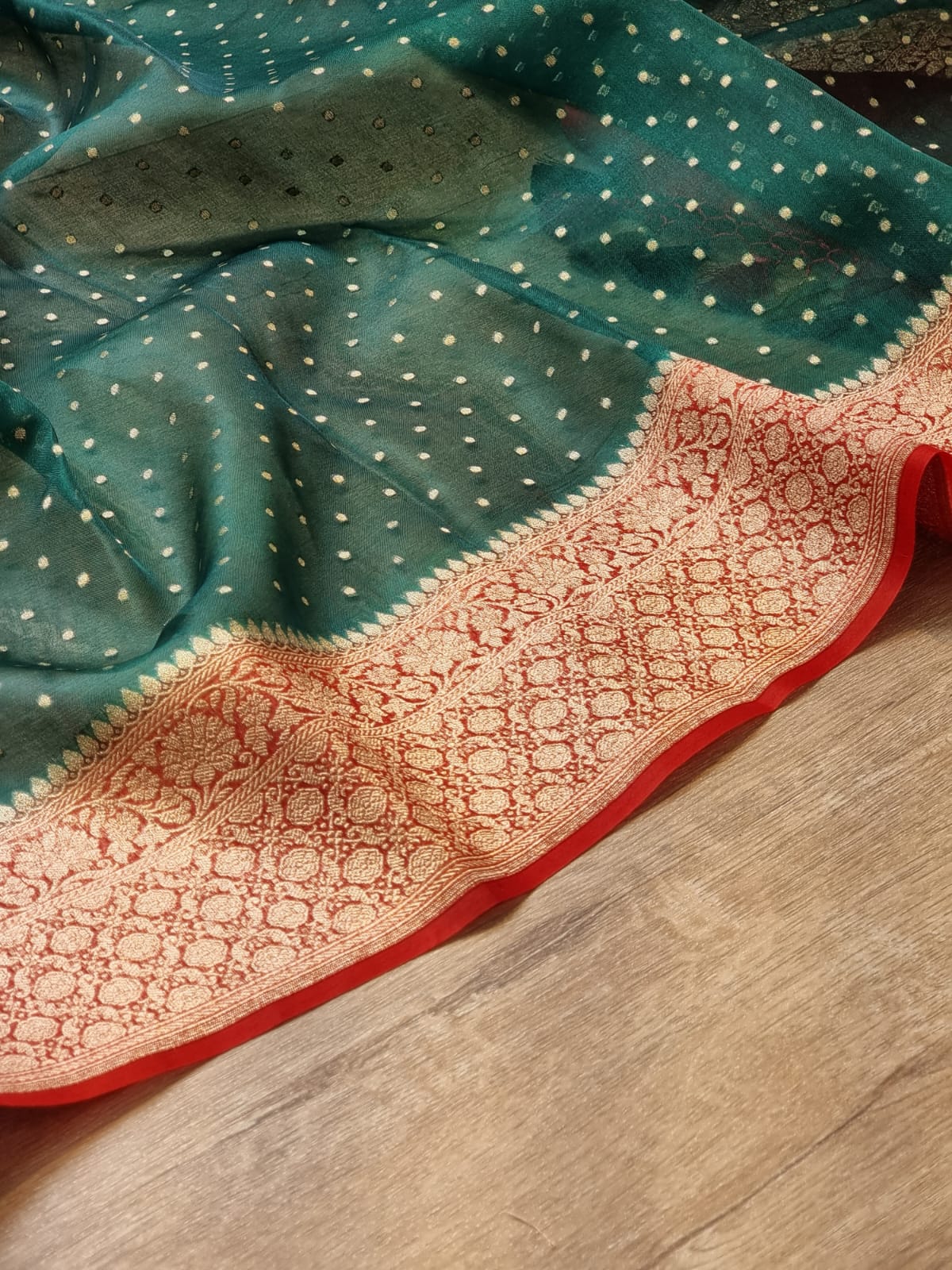 A teal green saree or fabric piece with small white dot motifs and a wide beige-gold brocade border, laid out on a light wood floor.