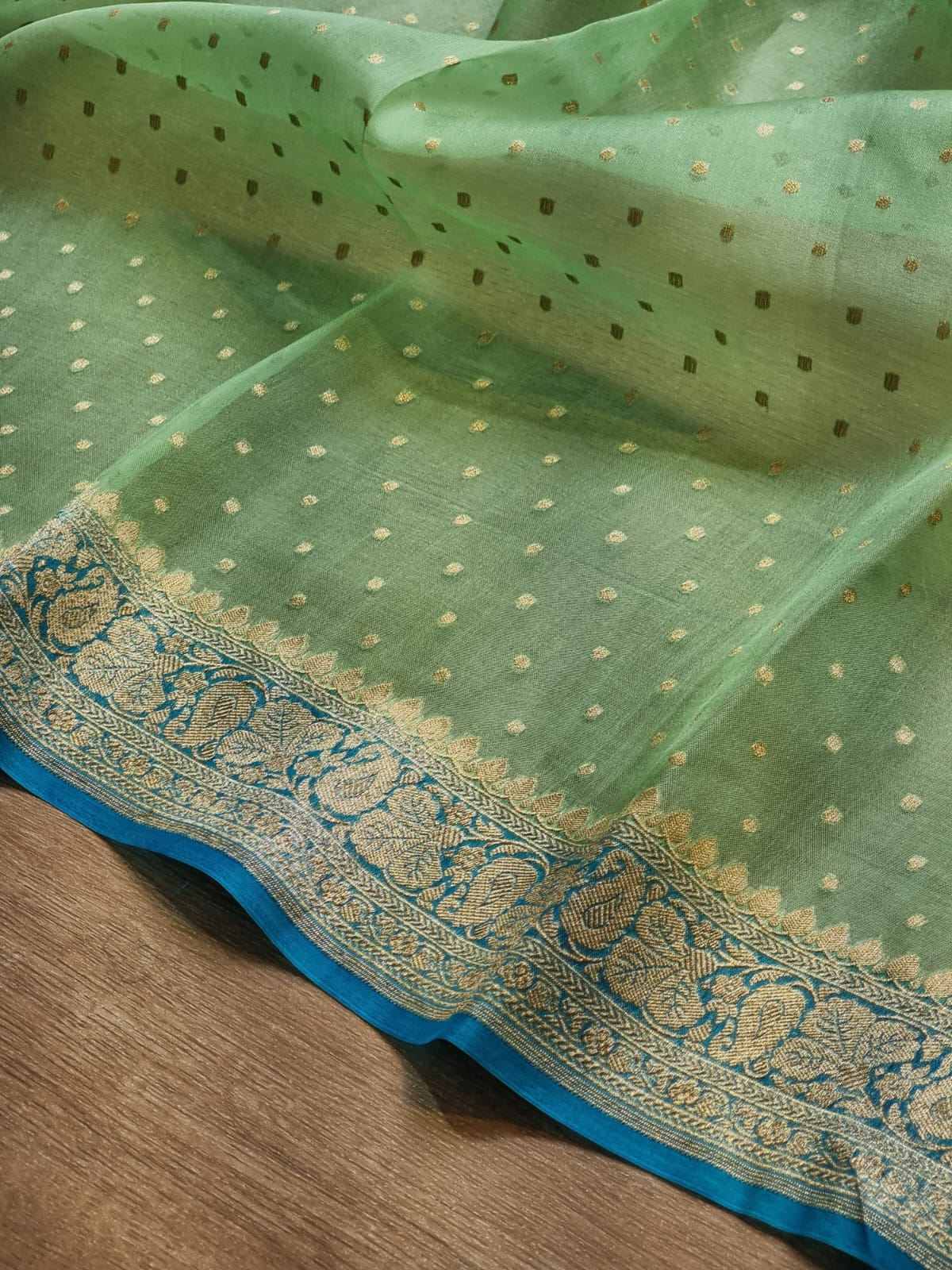 Light green sheer fabric, likely organza or silk, decorated with small gold and white embroidered dots and a wide blue and gold patterned border, laid on a wooden surface.